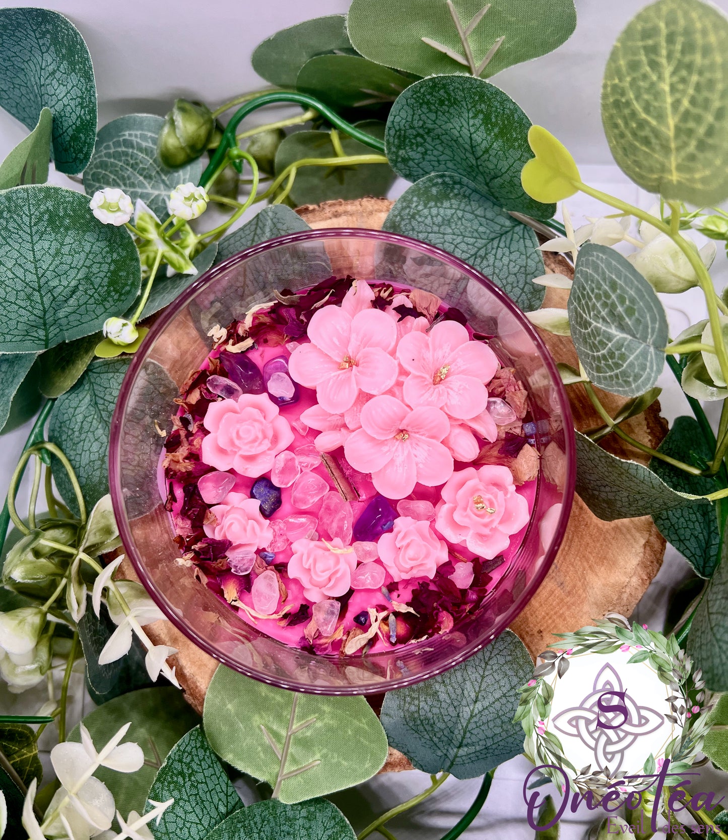 Bougie 'Féminin sacré' dans un contenant en verre rose, décorée de fleurs en cire et de pierres précieuses, posée sur un support en bois. Environnement naturel avec des feuilles vertes et des fleurs blanches.