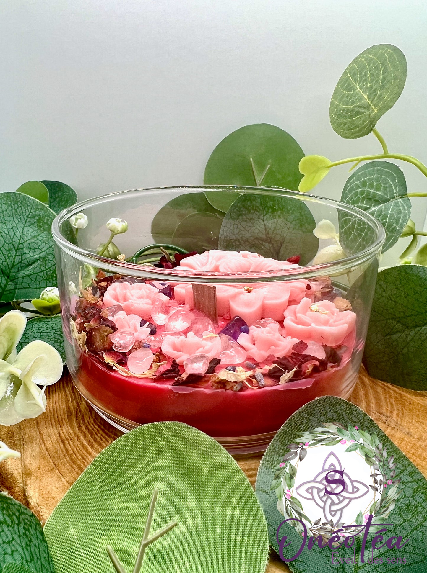 Bougie 'Féminin sacré' dans un élégant récipient en verre transparent, remplie de pétales et de pierres précieuses multicolores, entourée de feuillage vert. Un aspect serein et apaisant.