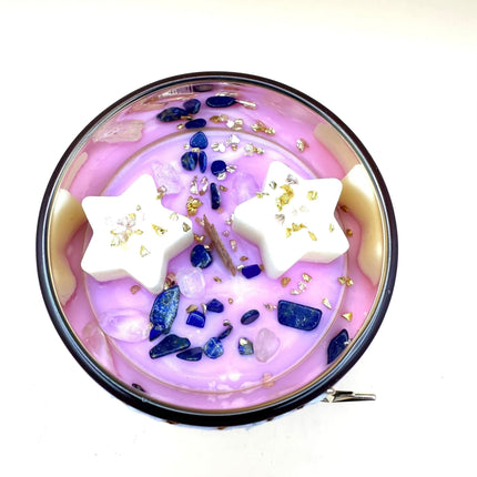 Bougie Résilience en verre à la cire rose et mauve, ornée de pierres bleues, d’éclats dorés et de deux étoiles blanches.