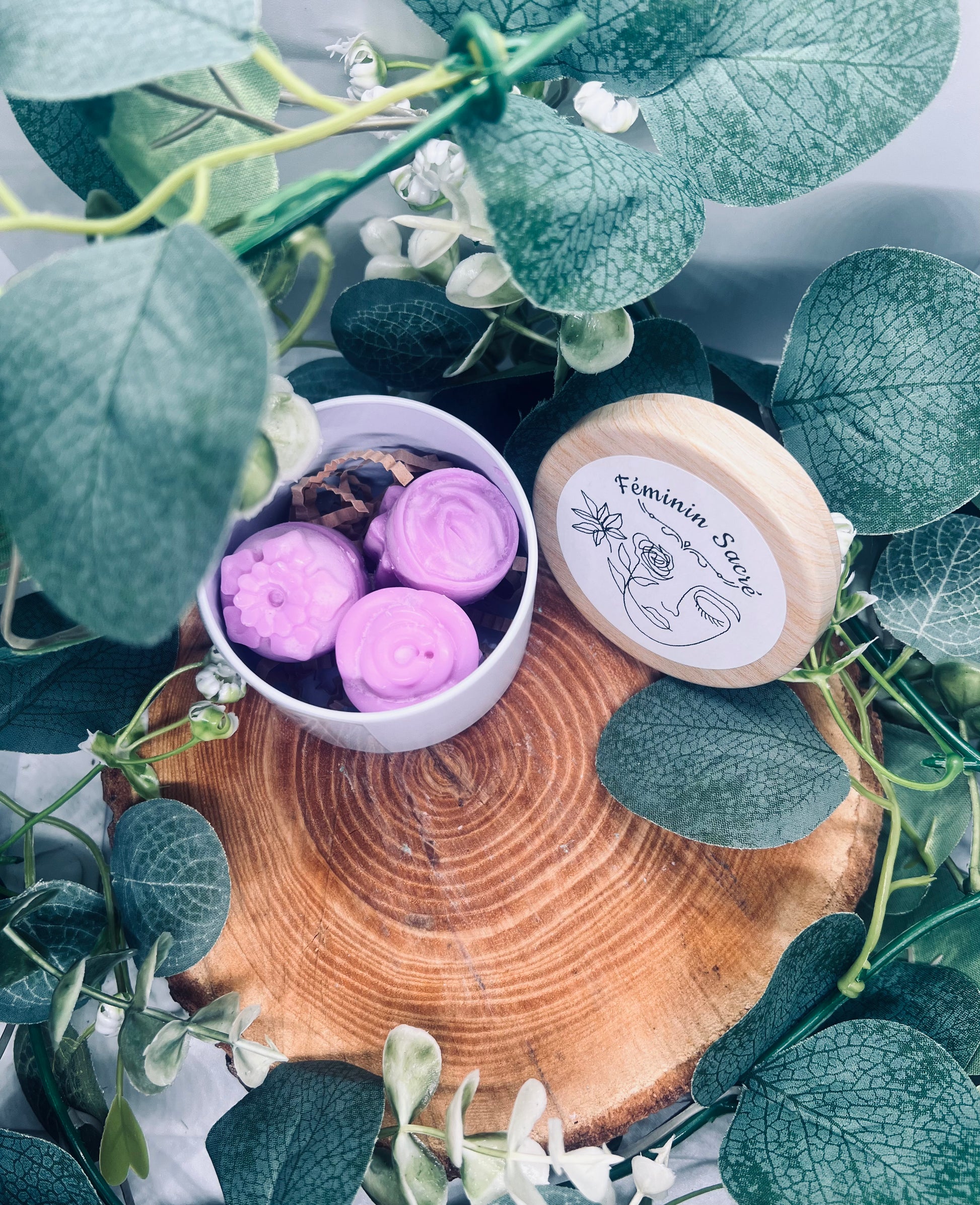 Un fondant parfumé 'Féminin sacré' dans un contenant en carton, entouré de feuilles vertes et disposé sur un fond en bois, pour une touche d'élégance naturelle.