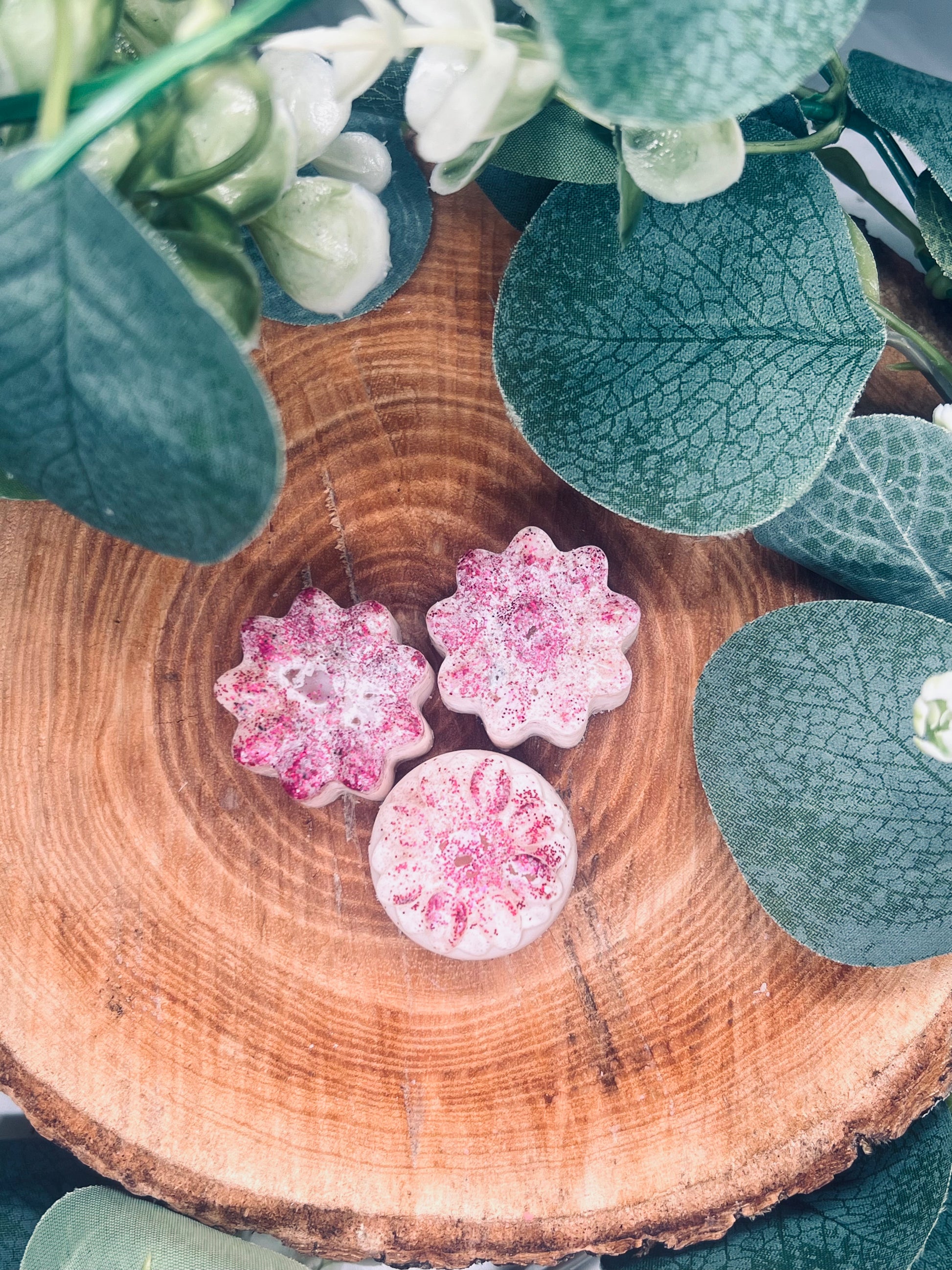 Trois fondants parfumés en forme de fleurs, ornés de paillettes roses, posés sur une rondelle de bois sous un feuillage verdoyant. Préparez votre esprit pour une aventure olfactive !