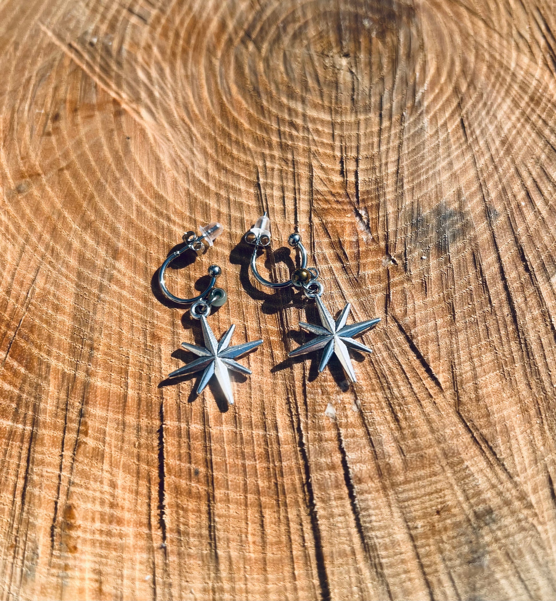 Créoles en acier inoxydable avec un pendentif en forme d'étoile à huit branches, posées sur un fond en bois rugueux.