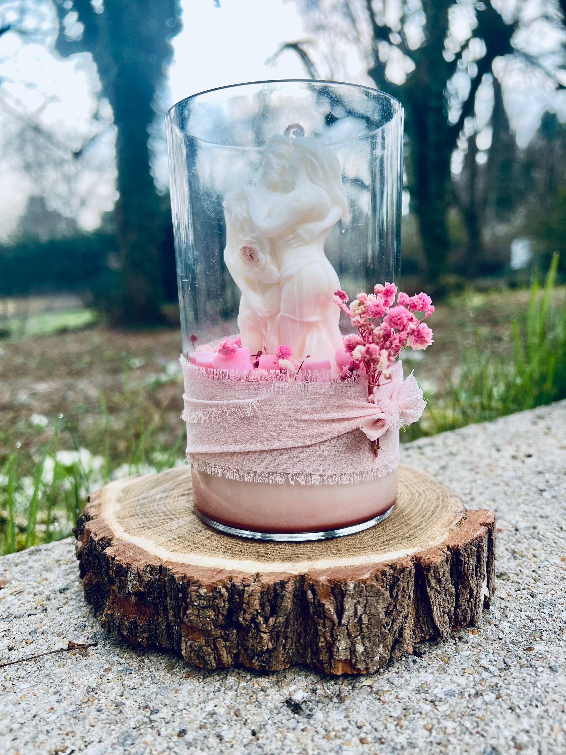 Vue latérale d'une bougie en verre représentant un couple, entourée de fleurs séchées roses, avec une décoration en tissu, sur une base en bois.