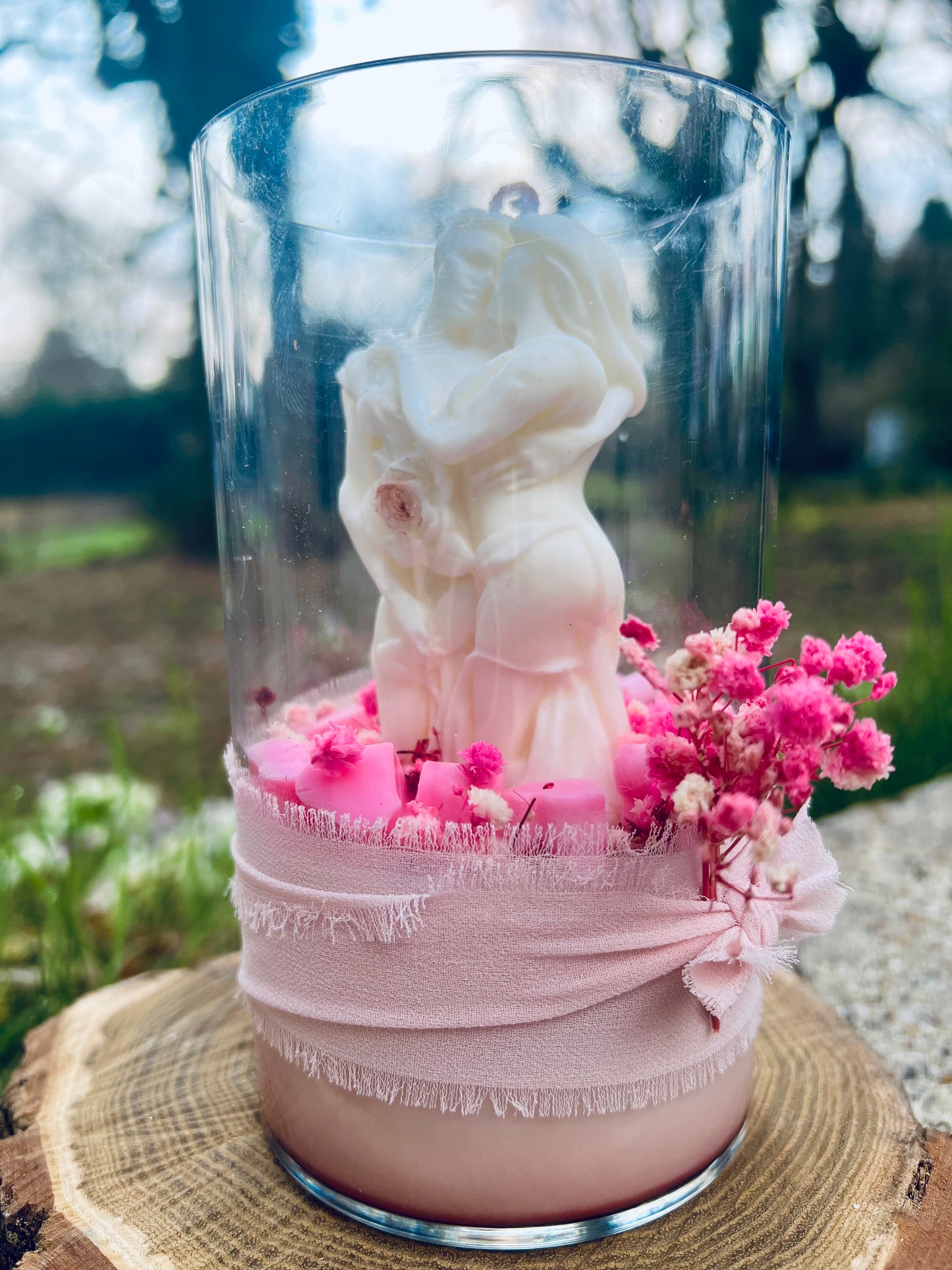 Bougie en verre décorée d'une figurine de couple et de fleurs roses, reposant sur une rondelle en bois, avec un fond flou de nature.