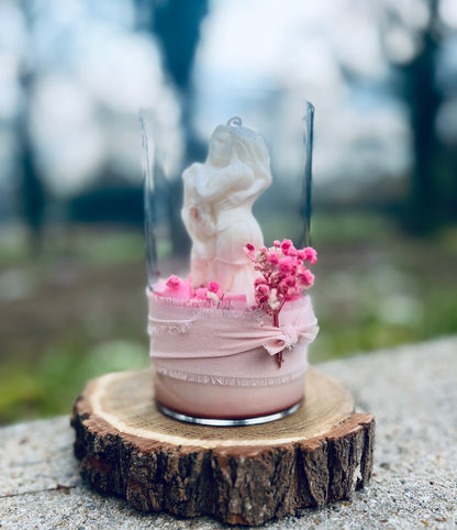 Bougie en verre avec une figurine de couple, ornée de fleurs séchées roses et un ruban en tissu, posée sur une tranche de bois.