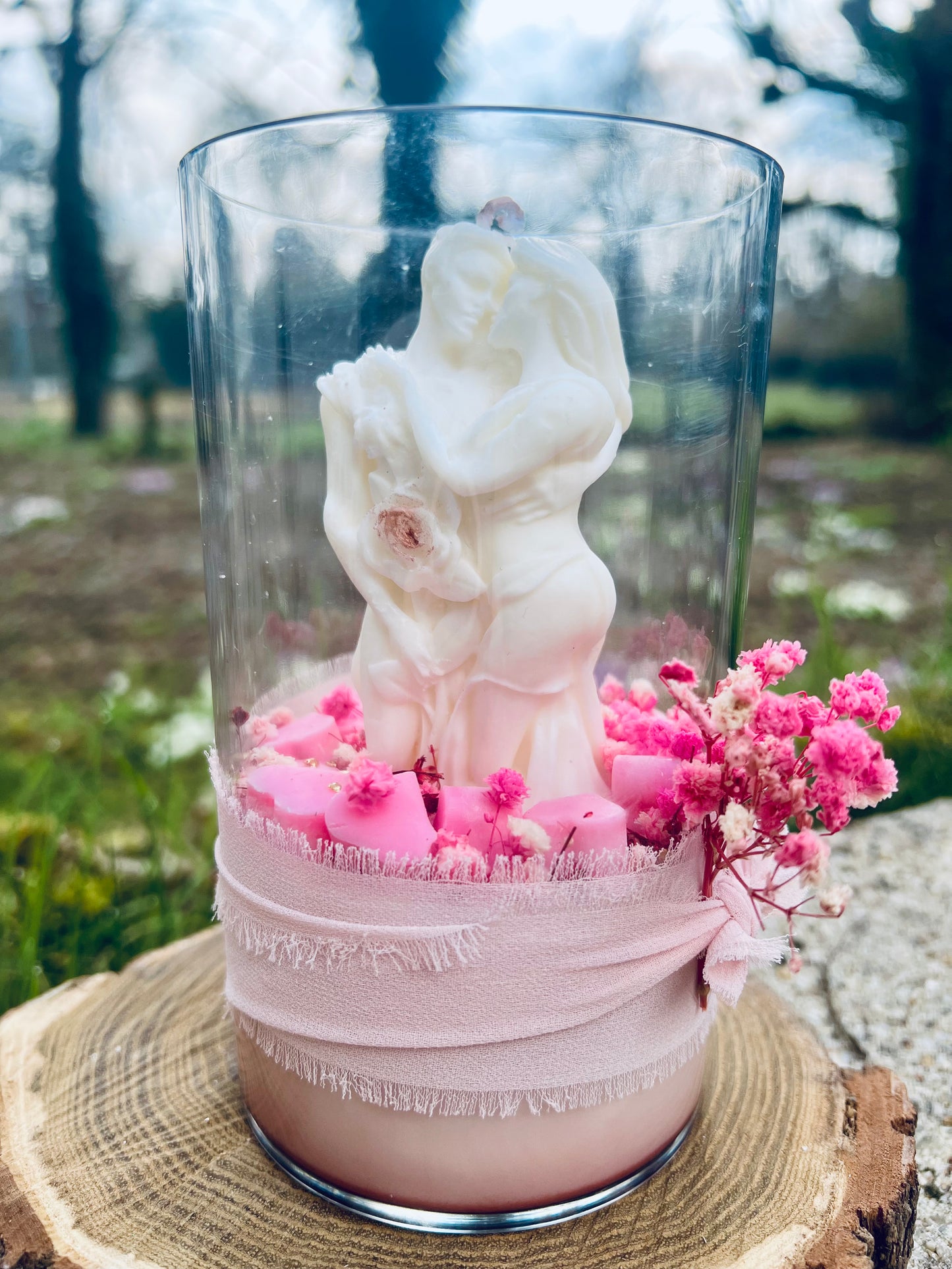 Bougie en verre avec figurine de couple, garnie de fleurs et un ruban, sur une base en bois, capturée en extérieur.