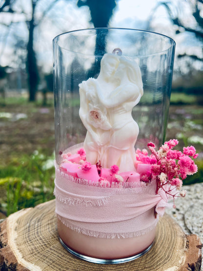 Bougie en verre sur une tranche de bois, ornée d'une figurine de couple et de décorations florales, dans un cadre naturel.