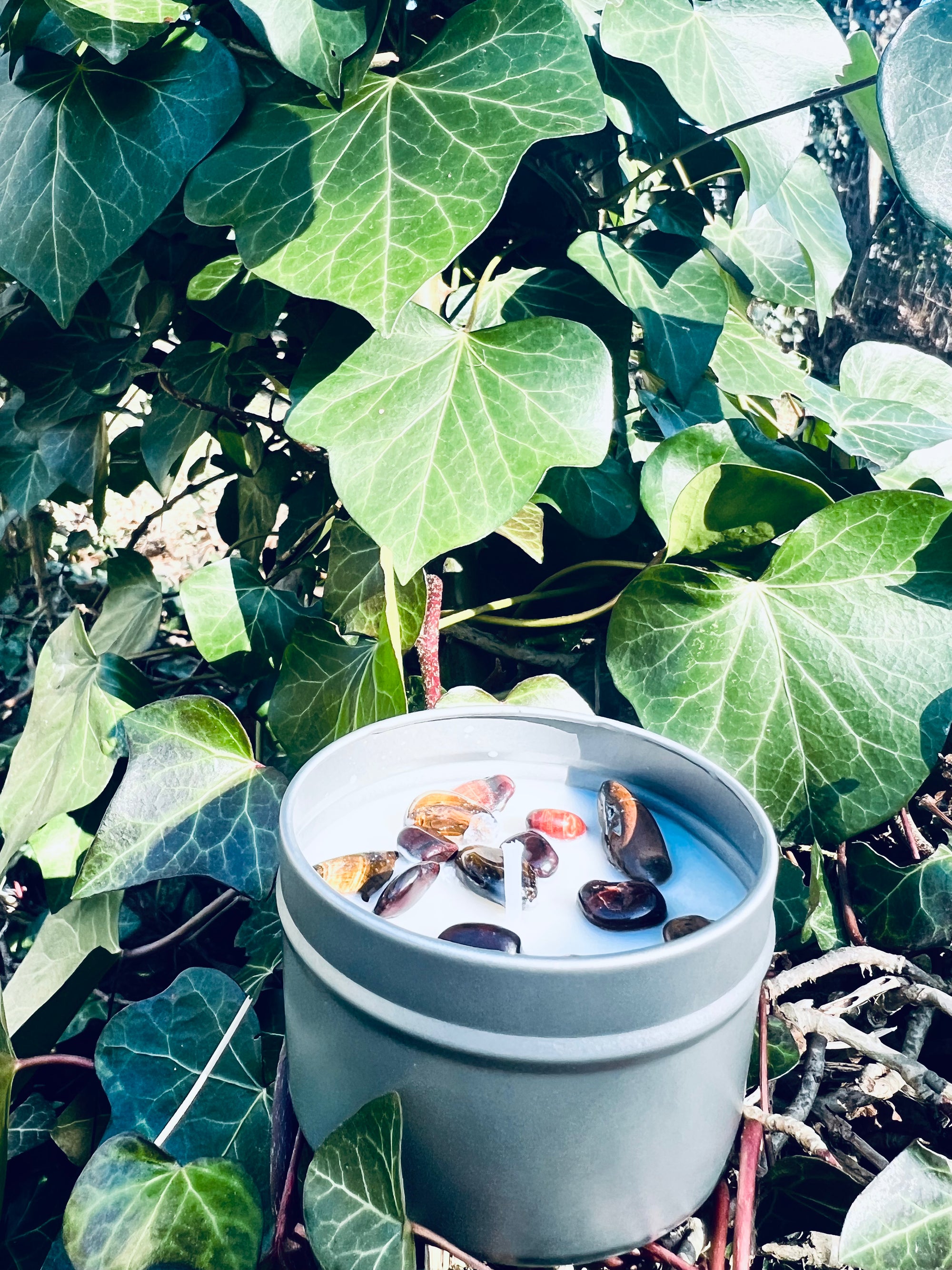 Bougie parfumée 'Oeil du Tigre' de Onéotéa - Éveil des Sens, entourée de verdure luxuriante avec une vue rapprochée sur des pierres symboliques, évoquant une atmosphère de protection et de bien-être.