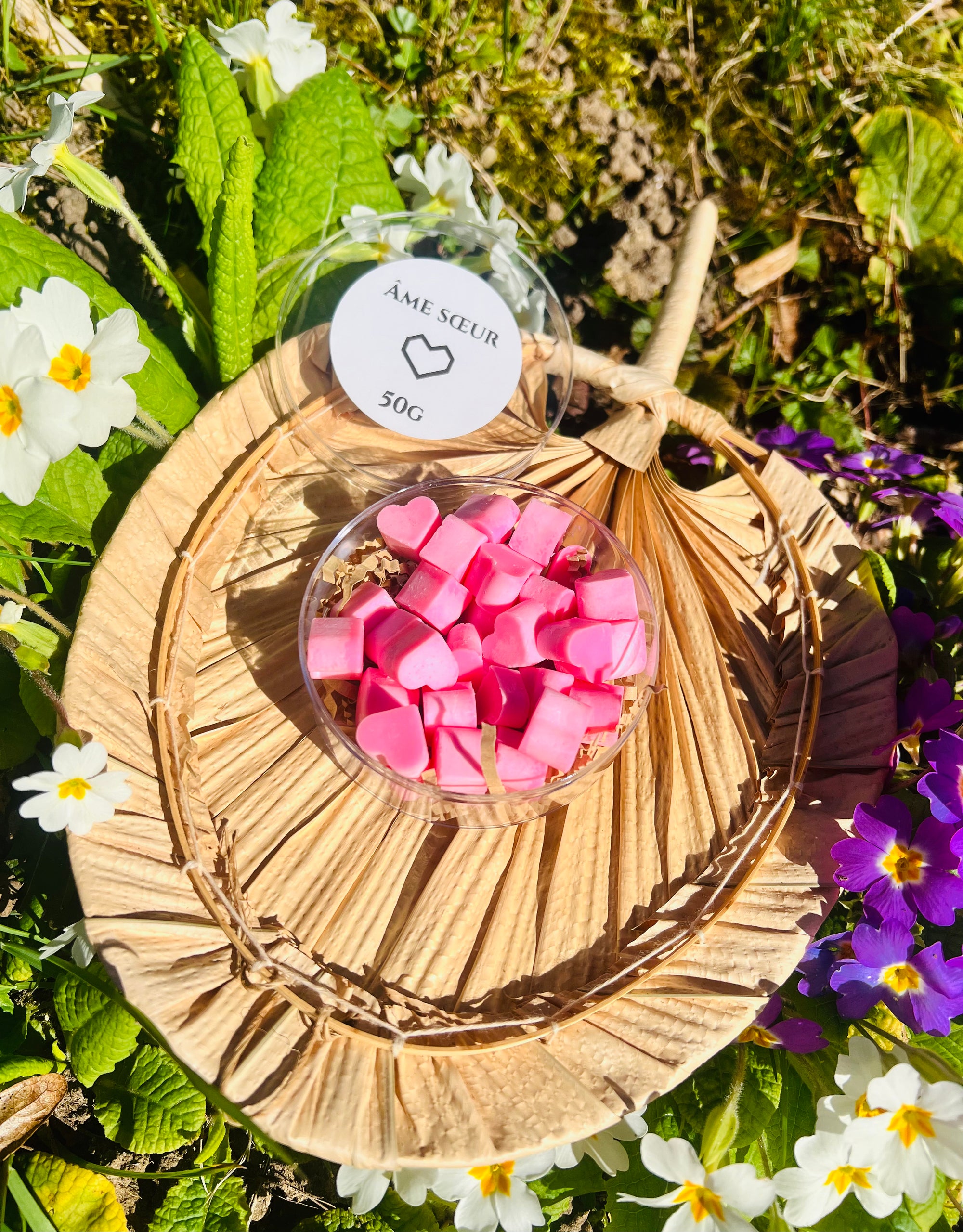 Vue rapprochée des délicats fondants parfumés 'Âme Soeur' d'Onéotéa - Éveil des Sens, dans un pot transparent sur un support naturel, avec des fleurs à proximité. Fondant parfumé, âme soeur.