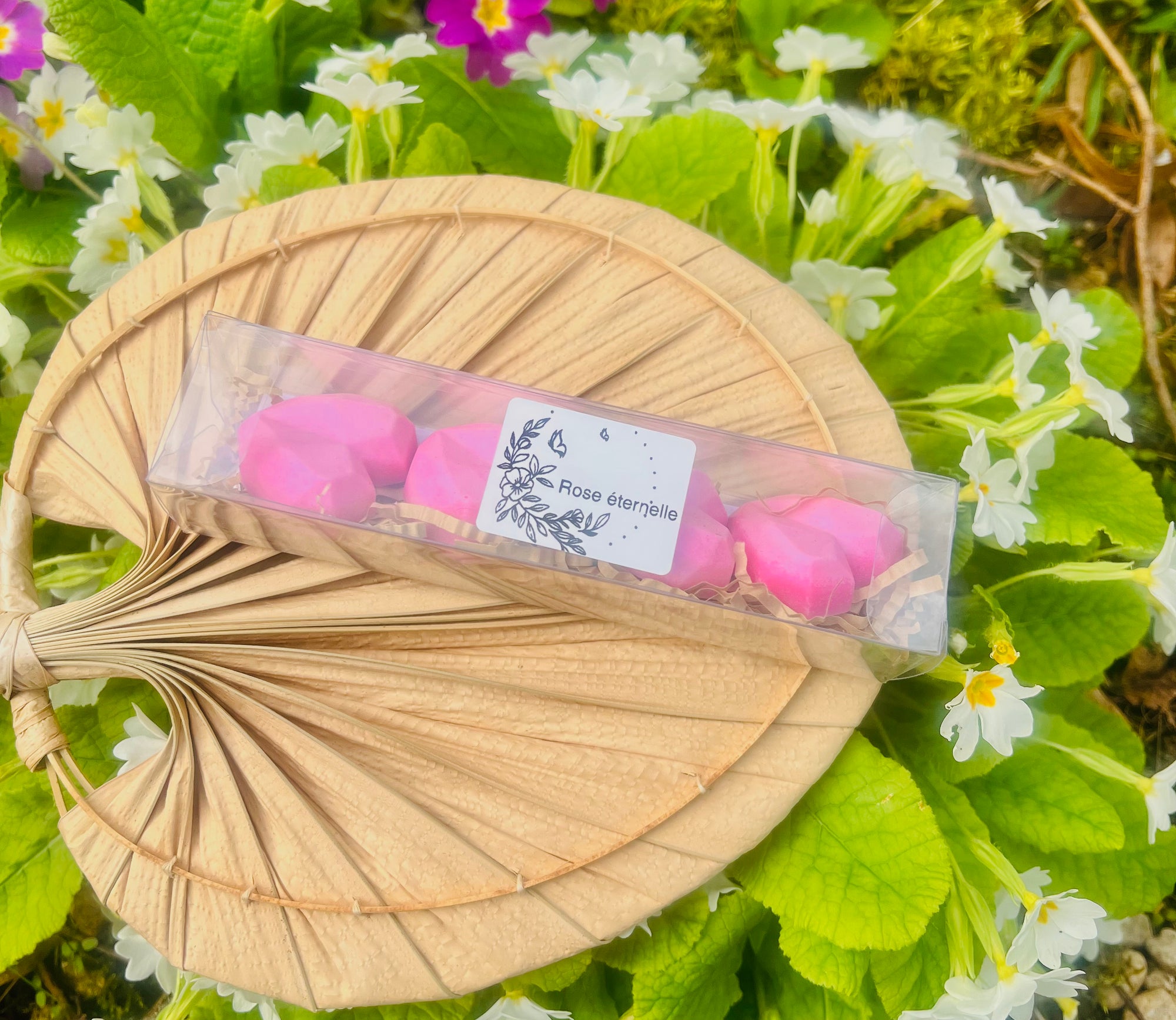 Fondants parfumés Rose éternelle de Onéotéa - Éveil des Sens, exposés dans leur boîte en plastique sur un éventail tressé, entourés de fleurs, soulignant la délicatesse de ces fondants en cire de soja au parfum de Grasse.