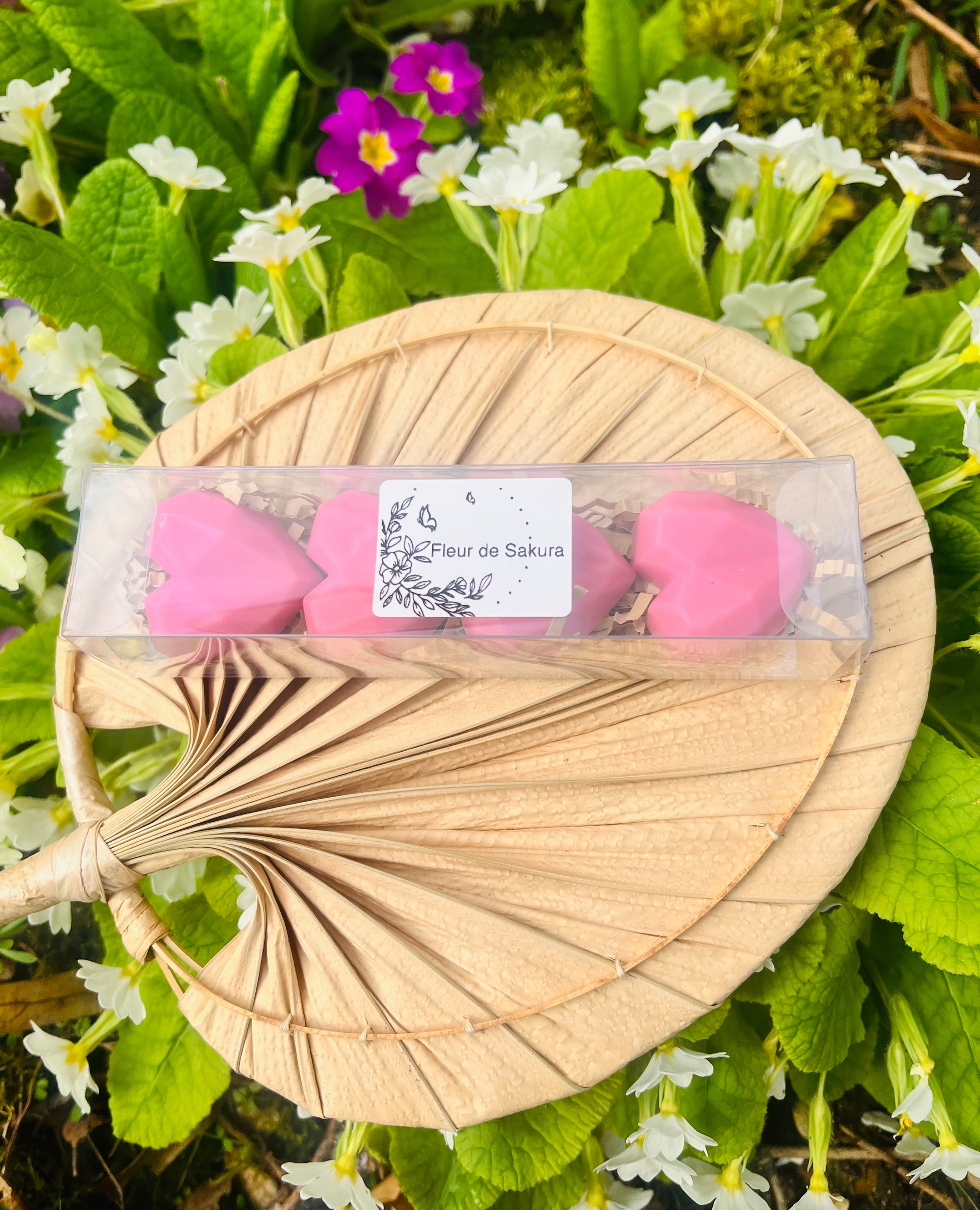 Boîte contenant des fondants parfumés en forme de cœur, de couleur rose, présentés sur un éventail en arrière-plan. Ces fondants parfumés de Onéotéa - Éveil des Sens, intitulés 'Fleur de SAKURA', sont fabriqués à partir de cire de soja et contiennent un parfum de Grasse. Fondant parfumé, fleur de Sakura, cire de soja, parfum de Grasse.