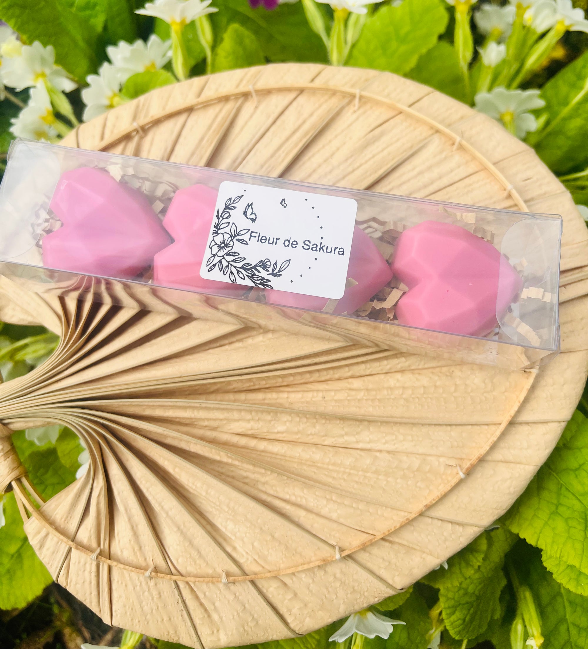 Vue rapprochée de la boîte de fondants parfumés en forme de cœur, rose, sur un éventail naturel. Ces produits de Onéotéa - Éveil des Sens, baptisés 'Fleur de SAKURA', allient délicatesse et fragrance, avec une composition de cire de soja et parfum de Grasse. Fondant parfumé, fleur de Sakura, cire de soja, parfum de Grasse.