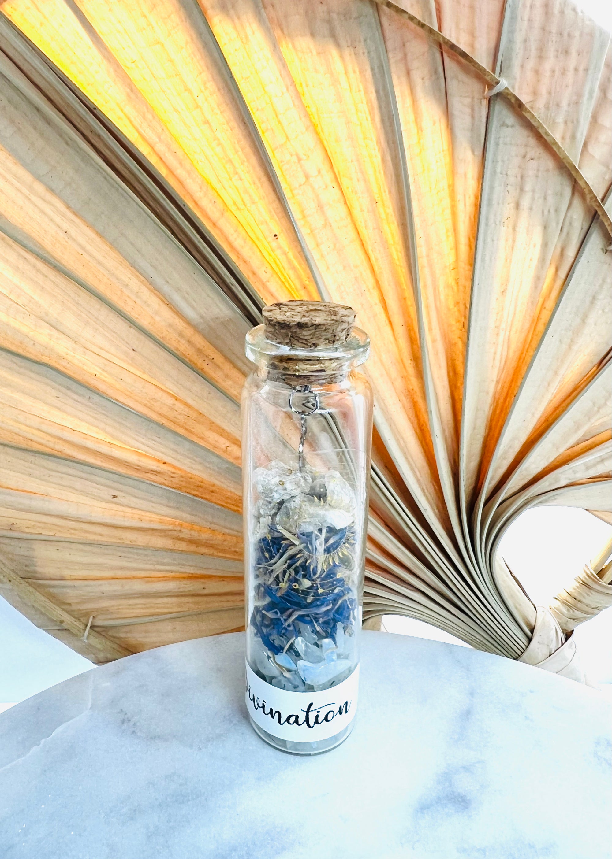 Fiole de divination en verre de Onéotéa, remplie de pierre de lune et de fleurs de bleuet, sur une table en marbre et un fond de palmiers, pour une touche magique.